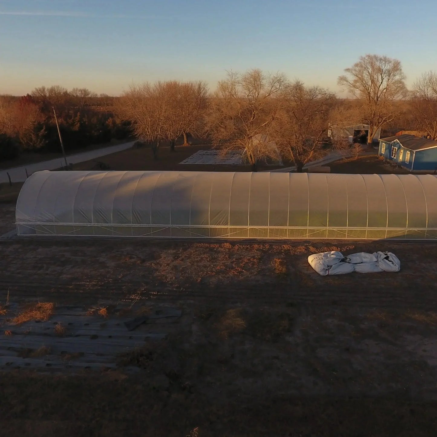 All-metal hoop house greenhouse on a rural farm at sunset, surrounded by trees and open land.
