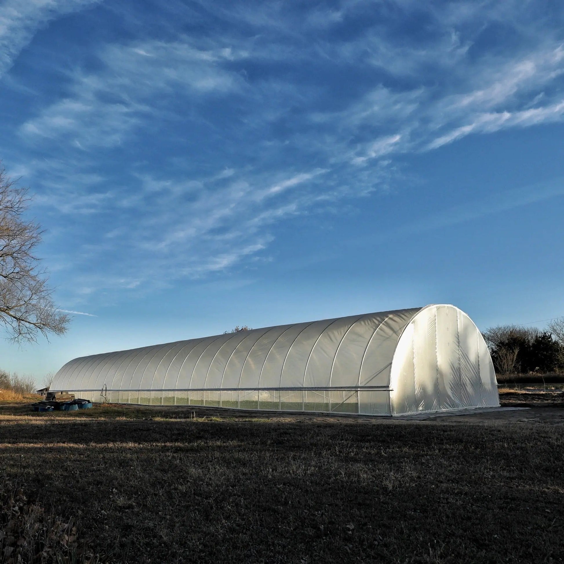 Large all-metal hoop house greenhouse in a rural farm setting under blue sky